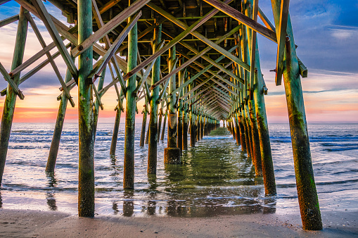 Light from a rising sun illuminates the pilings beneath the pier at Folly Beach, South, Carolina