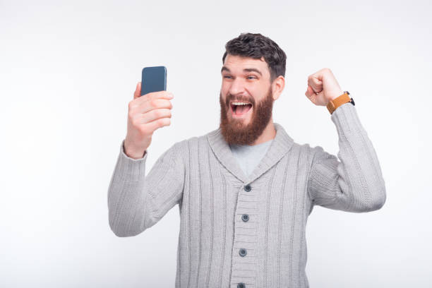 istock Young bearded man is making a winner gesture, looking at his phone and making a selfie. 1201282982