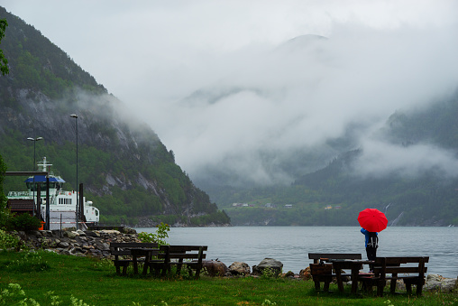 Female tourist with red umbrella on fjord shore, taking photo with camera. Tourism and traveling concept.