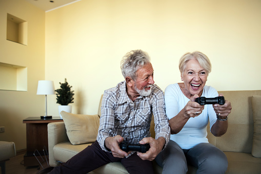An older couple with grey hair is playing video games at home. A senior couple is sitting on the sofa at home and holding gamepads while enjoying video games.