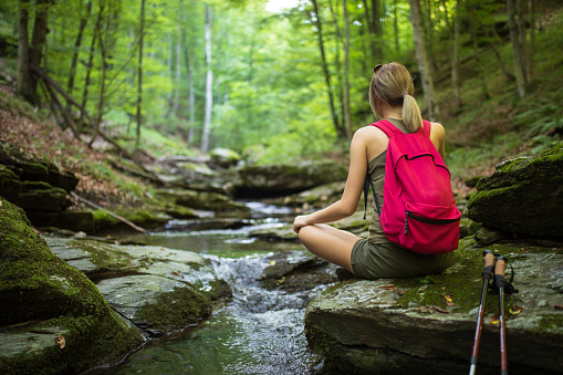Young woman with healthy habits hiking and relaxing in deep forest on mountain. Small stream flowing through this amazing place. She is alone, but very confident. She wears green shorts and t-shirt and has a red backpack, hiking boots and poles