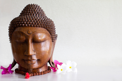 A carved wooden Buddha on a white background, with mala beads and flowers. Wellness/ yoga/ meditation stock photo.