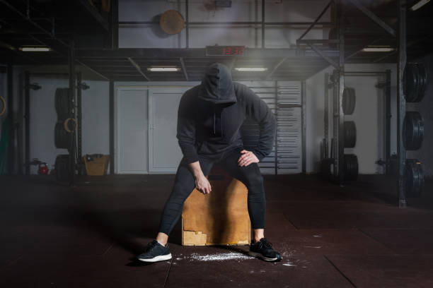 Young fit man in black sweatsuit with hood on his head sitting on the jump box in the gym taking a rest after hard core cross workout training Taking a break, Young fit man in black sweatsuit with hood on his head sitting on the jump box in the gym taking a rest after hard core cross workout training crossfit chalk weightlifting weight training stock pictures, royalty-free photos & images