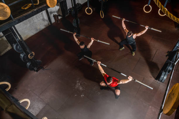 grupo de três jovens amigos se encaixam em caras de fitness muscular se divertindo por treino de treino cruzado duro na academia com barbells e agachamentos visão de foco seletivo de cima - crouching barbell weightlifting weight training - fotografias e filmes do acervo