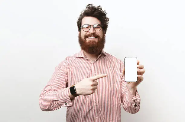 Portrait of happy bearded man with curly-hair pointing to white screen of his smartphone, wearing glasses