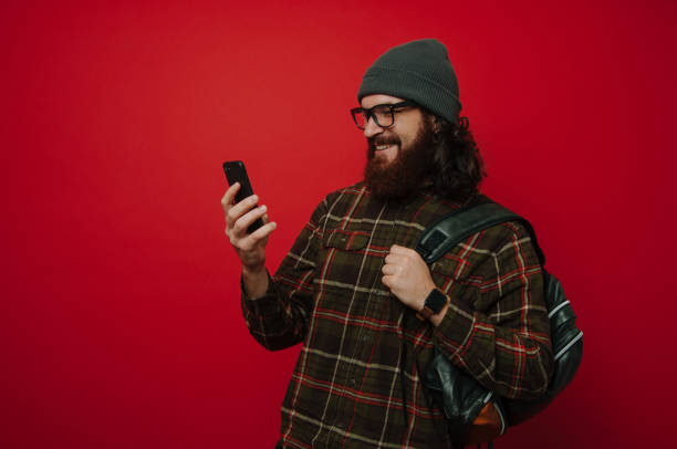 istock Portrait of smiling hipster man using a phone and holding backpack, over red background 1201300928