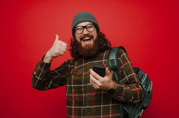 istock Happy  bearded man, using smartphone andgesturin with tumb up stanting over red background 1201301647
