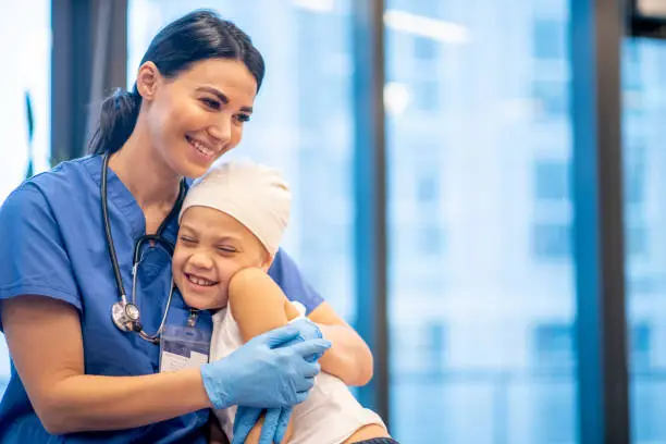 A young Latin female nurse hugs her cancer patient as they enjoy a brief moment together.  The young girl is smiling and happy.  The nurse is wearing blue scrubs with a stethoscope around her neck, while the young girl is dressed casually in a white tank top and has a head scarf on.