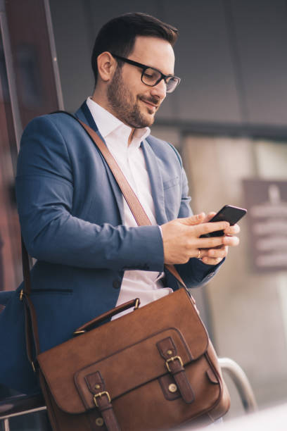 A young businessman outdoors, looking at his phone. He is carrying a bag over his shoulder. stock photo