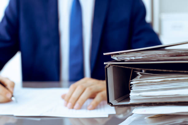 istock Binders with papers waiting to be processed with businessman or bookkeeper back in blur. Accounting planning budget. Audit, insurance and business concept 1201328086