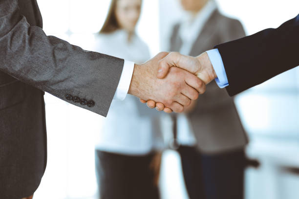 istock Business people shaking hands while standing with colleagues after meeting or negotiation, close-up. Group of unknown businessmen and women in modern office. Teamwork, partnership and handshake concept, toned picture 1201328186