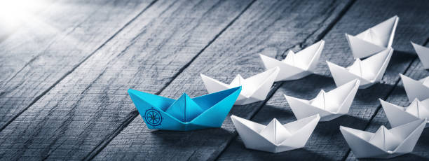 Blue Paper Boat Leading A Fleet Of Small White Boats Blue Paper Boat Leading A Fleet Of Small White Boats With Compass Icon On Wooden Table With Sunlight - Leadership Concept lead photos stock pictures, royalty-free photos & images