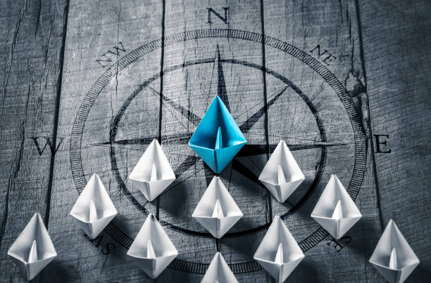 Blue Paper Boat Leading A Fleet Of Small White Boats Blue Paper Boat Leading A Fleet Of Small White Boats With Compass Icon On Wooden Table - Leadership Concept role model stock pictures, royalty-free photos & images