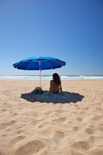 back brunette leisure rest woman in bikini sit on towel shade under blue parasol or sun umbrella sunshade summer holidays in sand beach with far water ocean sea near Conil de la Frontera Cadiz Andalusia Spain Europe