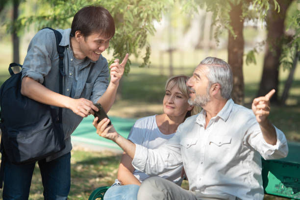 彼の手にスマートフォンを持つアジアの観光客の若者は、女性と一緒に座っている古い白人の長老の男性からの道順を求めます, 彼は道に指を向けた. - 問う 写真 ストックフォトと画像