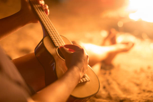 una donna musicista che suona la chitarra ukulele accanto a un falò sulla spiaggia. - hawaiian food foto e immagini stock