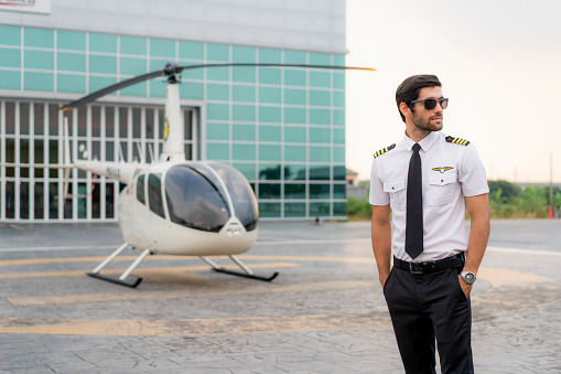 Portrait of handsome commercial pilot in captain white uniform standing and looking smart near small private helicopter on a landing point with private jet terminal at international airport in background.