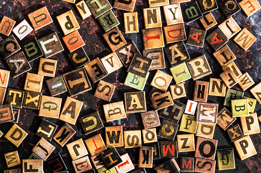 Color image depicting an overhead view of a large selection of colored alphabet letter tiles scattered, apparently haphazardly, across a rusty metal surface.