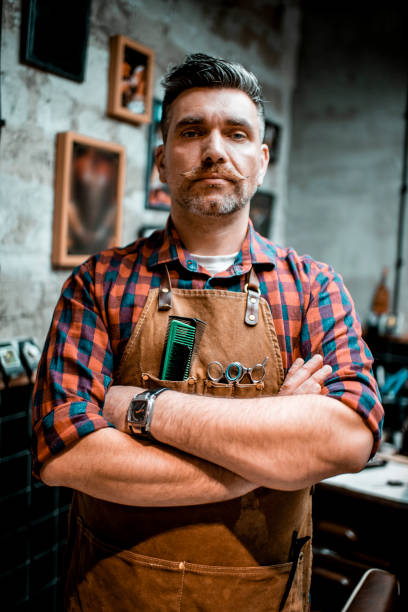 istock A barber with twirled mustache and an apron posing for camera in a barber shop 1201401418