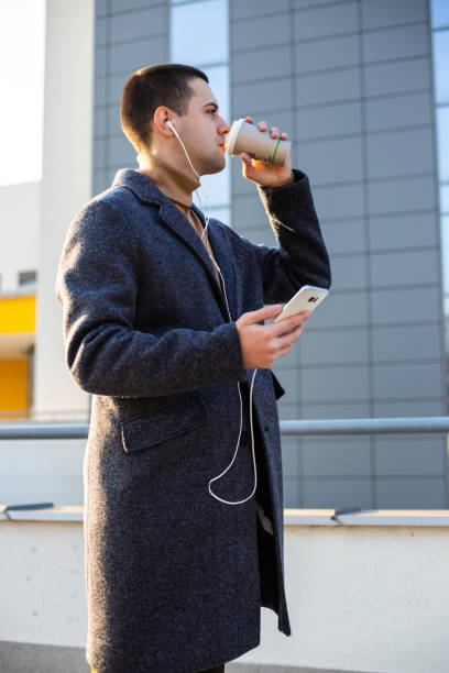 istock Elegant young man is drinking coffee to go and using smartphone outside 1201403046