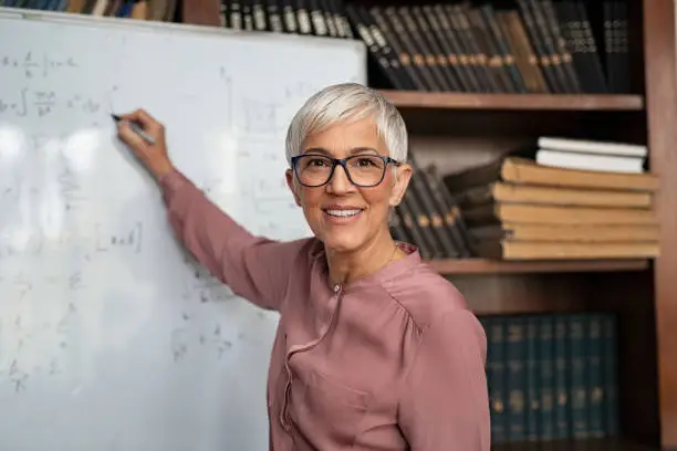 Portrait of happy mature professor teaching mathematics to students in a library. Senior smiling woman solving math problem while writing on white board with a marker. Portrait of tutor looking at camera.