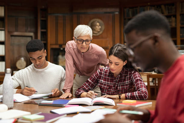 University students studying with teacher Senior woman teacher working with college students in library. Graduates consulting with their lecturer informally during a break in library. Group of students studying with their professor. librarian stock pictures, royalty-free photos & images