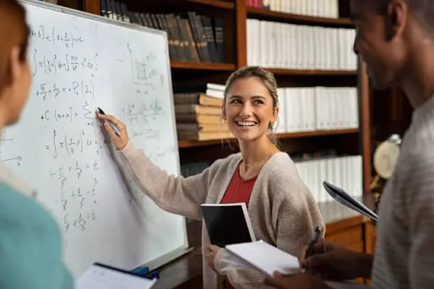 Happy girl writing math formulas on whiteboard while looking her classmate. Young woman solving arithmetic problem while standing with university students in classroom. Smiling college student holding book explaining math problem at school.