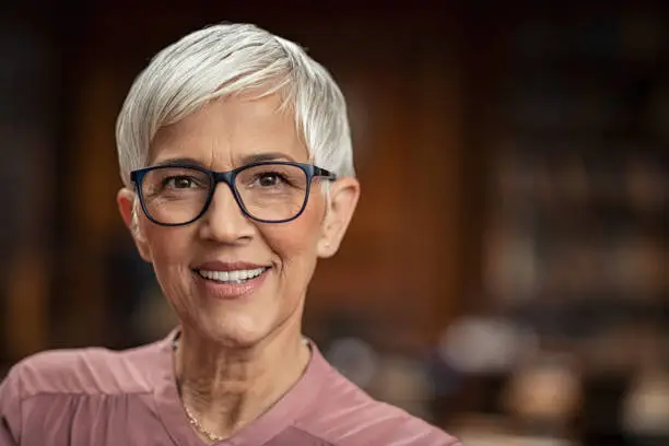 Portrait of beautiful senior woman looking at camera with copy space. Successful mature business woman wearing eyeglasses. Happy old professor in standing in college library with gray hair.