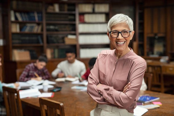 Smiling university professor in library Portrait of mature professor with crossed arms standing in university library and looking at camera with copy space. Happy senior woman at the library working as a librarian. Satisfied college teacher smiling with students in background studying. lecturer stock pictures, royalty-free photos & images