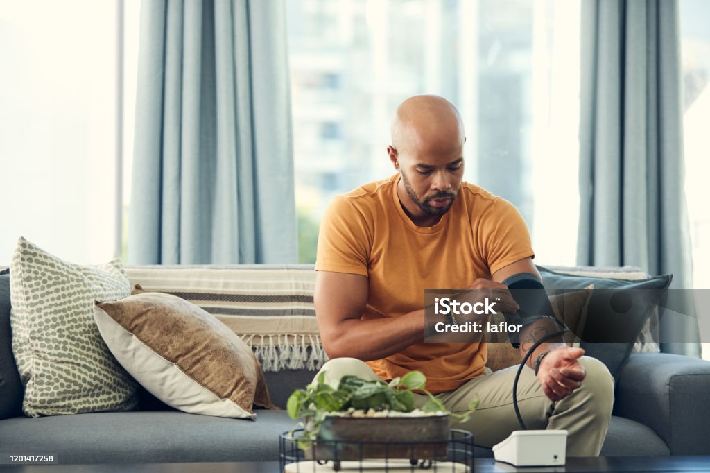 I'm experiencing headaches so my blood pressure might be high Shot of a young man taking his blood pressure while sitting on the sofa at home Blood Pressure Gauge Stock Photo