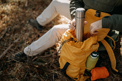 High angle view of mature man resting and taking insulated drink container from backpack while sitting in forest