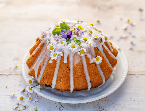 Easter yeast cake (Babka) covered with icing and decorated with edible flowers on a white plate on a white wooden table. Traditional Easter cake in Poland