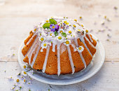 Easter yeast cake (Babka) covered with icing and decorated with edible flowers on a white plate on a white wooden table.