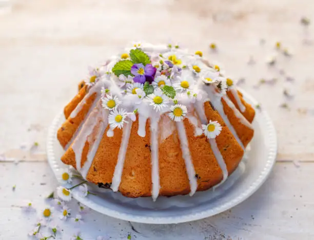 Photo of Easter yeast cake (Babka) covered with icing and decorated with edible flowers on a white plate on a white wooden table.
