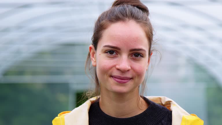 Close-up portrait of a young woman standing in plant nursery. Happy female gardener in raincoat looking at camera.