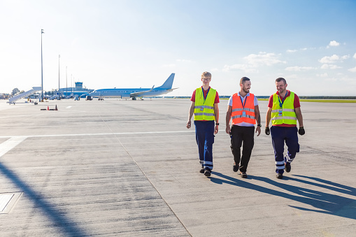 Three men walking on the airport runway with airplane in the background. Wide angle view.