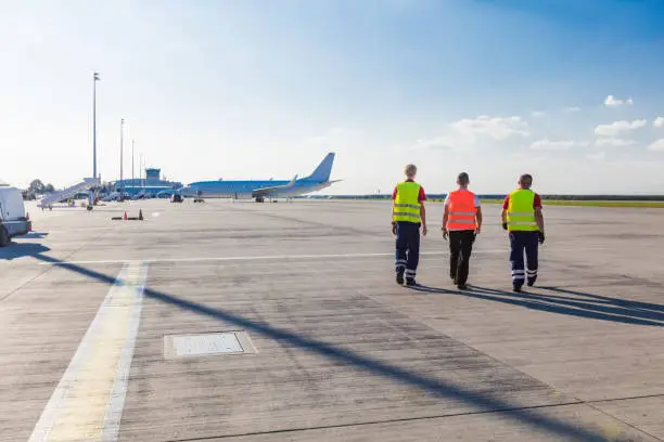 Rear view of three men walking on the airport runway with airplane in the background. Wide angle view.