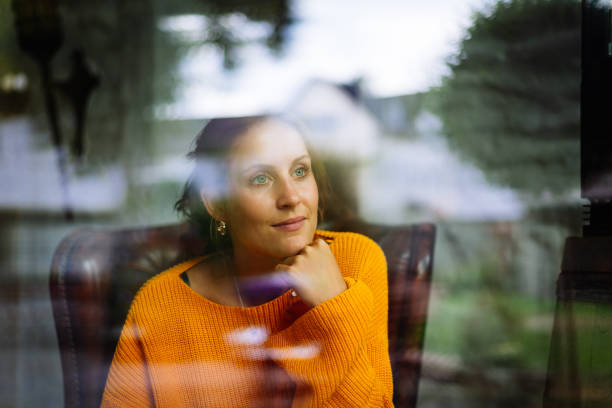 le jeune femme s'assied dans un fauteuil en cuir derrière une fenêtre et regarde dans l'appareil-photo - window reflection photos et images de collection