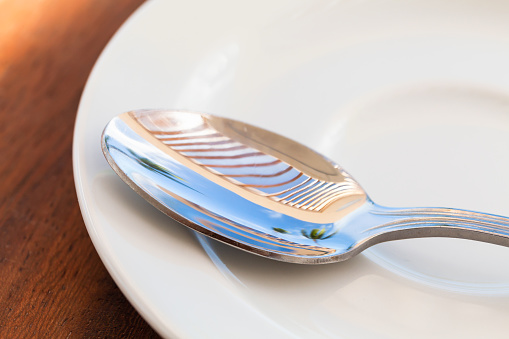 Shiny metal tea spoon with reflections of blue summer sky and palm trees lays on a white ceramic saucer, macro photo with selective focus