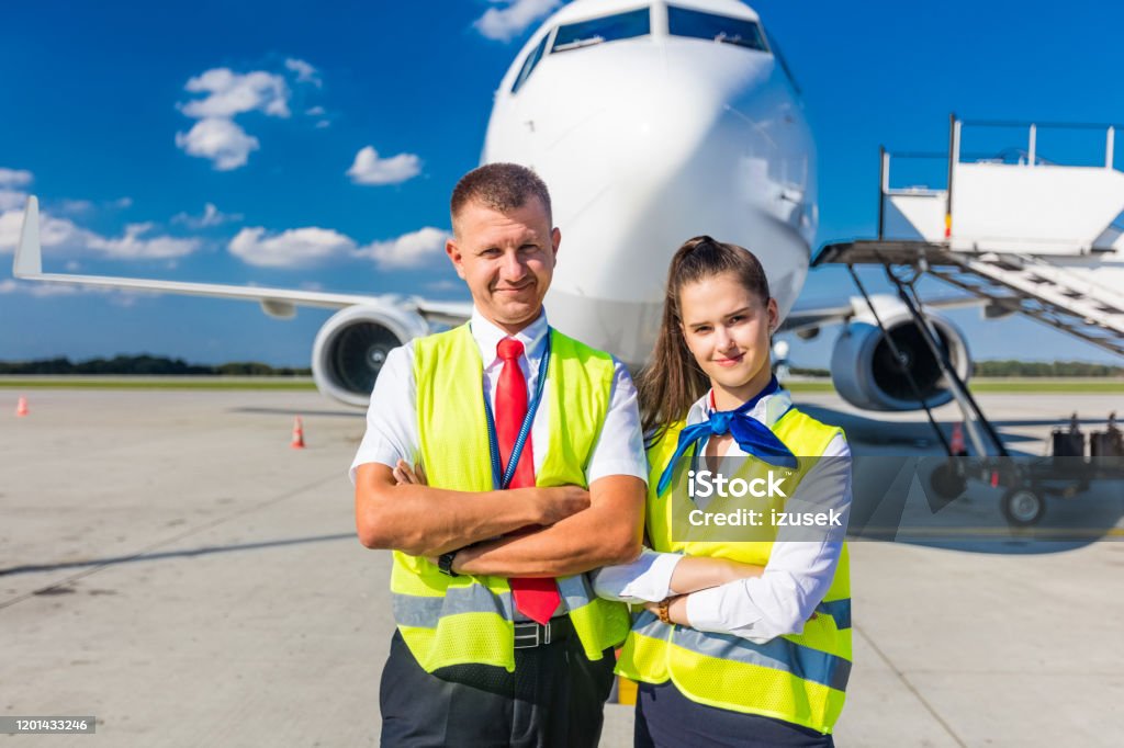 Airport service team in front of airplane Outdoor shot of young woman and man standing outside in front of airplane and smiling at camera. Airport ground crew at work. Airplane Stock Photo