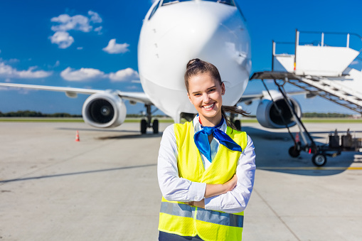 Outdoor shot of young woman standing outside in front of airplane and smiling at camera. Airport ground crew at work.