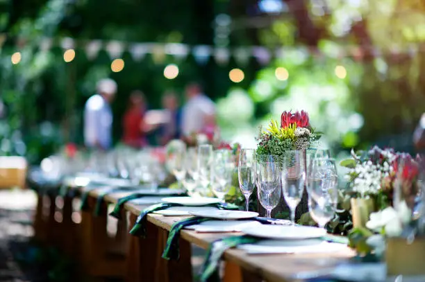 A protea on an outdoor wooden table set for a celebration in a forest during daytime with plates, glasses, napkins, fynbos flower arrangements and string lights and bunting defocussed with people behind Cape Town South Africa