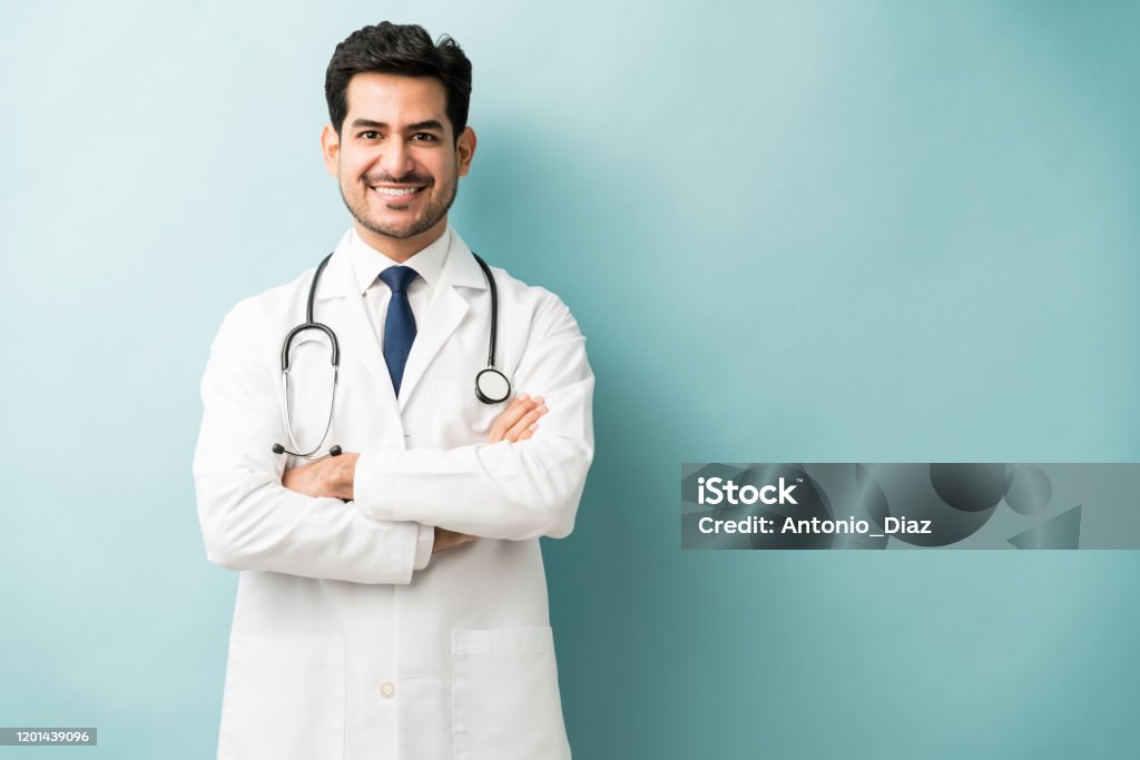 Male Medical Professional Is Confident In Studio Smiling Hispanic doctor standing with arms crossed against blue background Doctor Stock Photo