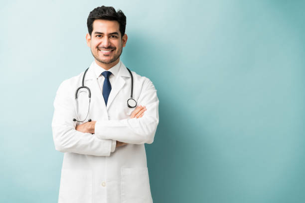 Male Medical Professional Is Confident In Studio Smiling Hispanic doctor standing with arms crossed against blue background necktie photos stock pictures, royalty-free photos & images