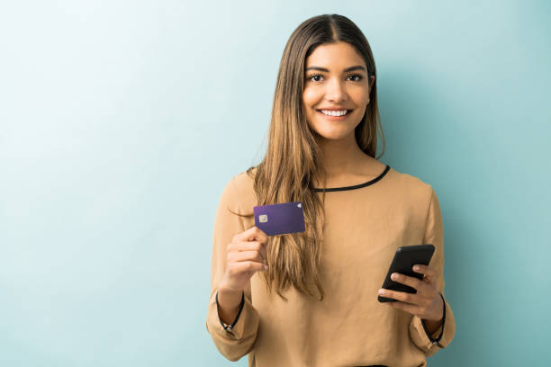 Happy Young Woman Doing Online Shopping In Studio Smiling Latin woman holding credit card and smartphone while standing against blue background young women shopping stock pictures, royalty-free photos & images
