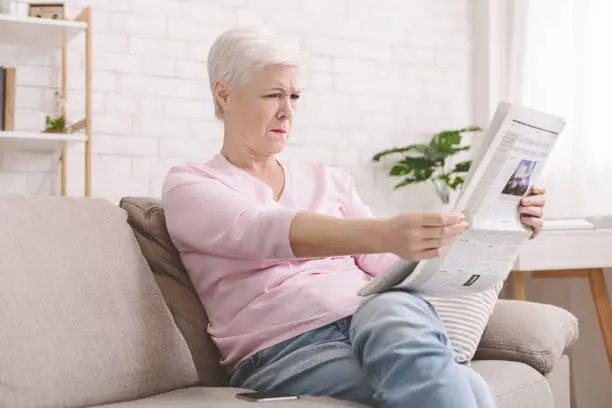 Bad sight problem. Senior lady squinting and holding newspaper far from eyes at home, free space