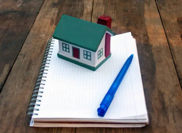 Photo of Squared notebook, small house and pen on an old table. Concept design, purchase, sale, rental, mortgage, mortgage, own house