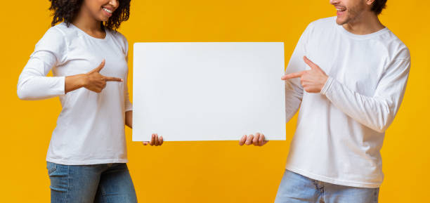 istock Interracial Couple Pointing At White Advertisement Placard In Their Hands 1201450439