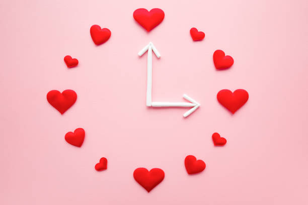 Clock created from bright red hearts and white arrows on light pink table background. Dating time. Love concept. Close up. Flat lay. Top down view.