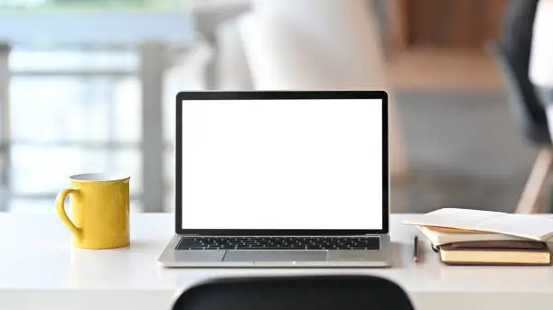 Photo of Laptop with white blank screen display and office equipment are putting on the white office desk over the modern office background.